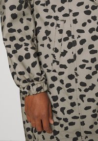 Nærbillede af en hånd ved siden af en grå stofærme med sorte uregelmæssige pletter og en lodret slidspose med synlige syninger.