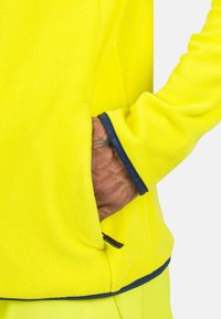 Pullover in pile di colore giallo brillante con dettagli in blu navy. Presenta un tessuto testurizzato, una tasca laterale e una bordatura a contrasto sui polsini.