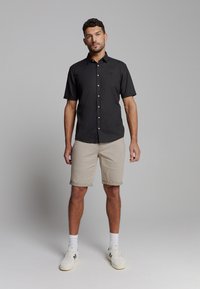 Zwarte korte mouwen button-up shirt, kaki shorts, witte sokken en sneakers; zachte stof, relaxte pasvorm en een neutraal kleurenpalet.