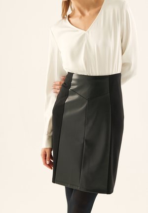 Kobieta w białej bluzce z długim rękawem i dekoltem w serek, wpuszczonej w czarną, skórzaną, ołówkową spódnicę z wysokim stanem, stojąca z jedną ręką na biodrze.