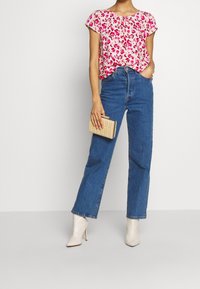 Femme portant un chemisier floral rose, un jean droit bleu, des bottes blanches à talons, et tenant une petite pochette beige.