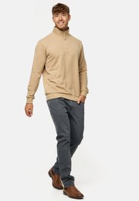 Sweatshirt beige à col zippé quart avec des manches longues, associé à un jean gris foncé et des bottes brunes. Le tissu semble doux et texturé.
