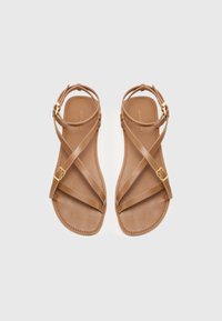 Bruine leren sandalen met een vlakke zool, voorzien van kruislings gekruiste bandjes en een gouden gespaccent op de enkelband. Glad textuur, minimalistisch ontwerp.