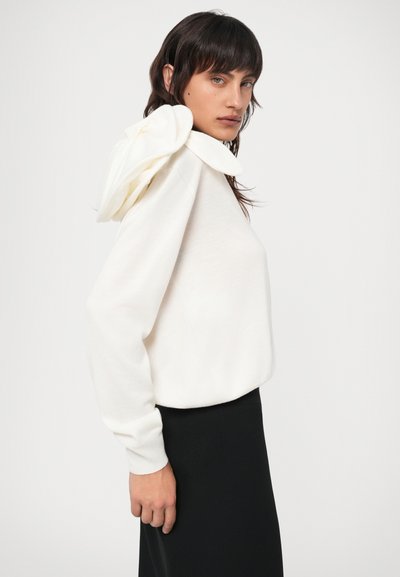 Balts džemperis ar unikālu kraukšķīgu kapuci, garām piedurknēm un brīvu piegriezumu; kombinēts ar melnu svārku kontrastam.