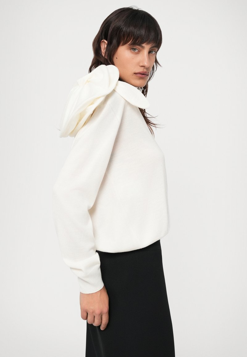 Felpa bianca con cappuccio arricciato unico, maniche lunghe e vestibilità rilassata; abbinata a una gonna nera per contrasto.