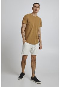 Brown short-sleeve T-shirt paired with light gray shorts. Model wears black sneakers. Tattoos visible on arms. Simple, casual design.