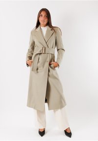 Cappotto trench beige doppiopetto con una cintura larga e tasche; presenta un colletto strutturato e un tessuto liscio, abbinato a pantaloni crema e tacchi neri.