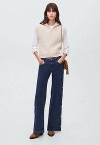 Top smanicato beige in maglia con colletto zip, abbinato a jeans svasati blu scuro con bottoni laterali. Stivali alla caviglia marroni completano il look.