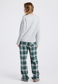 Femme de dos portant un pull gris, un pantalon à carreaux verts et blancs, et des pantoufles beiges sur un fond clair et uni.