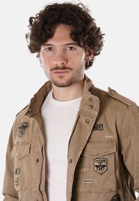 Giacca khaki con toppe e dettagli ricamati, dotata di chiusura con zip, tasche e tessuto testurizzato. Indossata sopra una maglietta bianca.