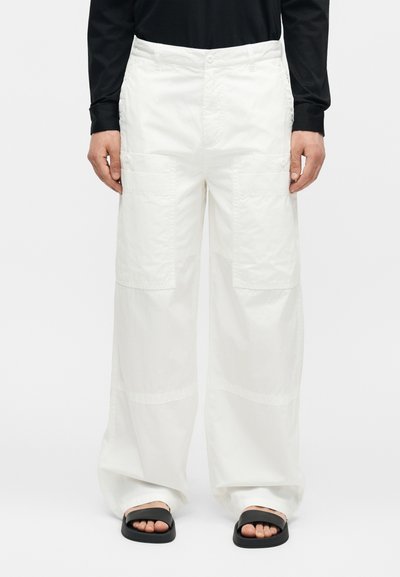 Personne portant un pantalon cargo blanc ample avec de grandes poches avant, un t-shirt noir à manches longues et des sandales noires à enfiler sur fond blanc.