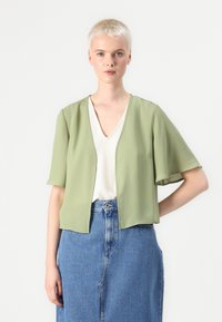 Lys grøn, kortermet blazer, V-halsdesign, myk tekstur, åpen front, lagd over en beige topp, sammen med et blått denimskjørt.