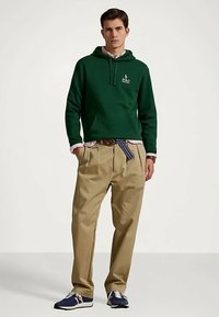 Ung man som bär en mörkgrön hoodie, beige veckade byxor med randig bälte och blå sneakers, står mot en enkel vit bakgrund.