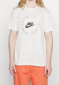 T-shirt en coton blanc avec un logo Nike noir et un design de texte circulaire en olive, à manches courtes et à coupe décontractée.