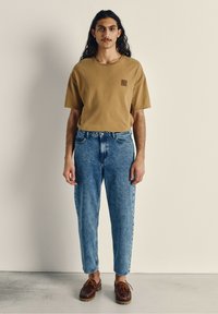 Brašna majica s kratkimi rokavi rjave barve z majhnim logotipom, v kombinaciji z bledosvetlimi oprijetimi jeans hlačami in rjavimi čipkastimi čevlji. Mehka tekstura in sproščen kroj.