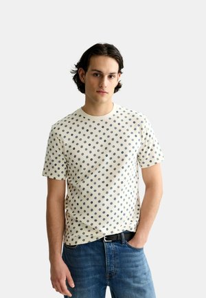 Junger Mann mit dunklen Haaren, der ein cremefarbenes T-Shirt mit kleinen blauen Sternmustern und blaue Jeans trägt, steht vor einem schlichten Hintergrund.