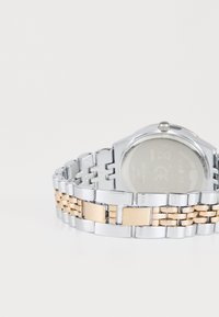 Montre en métal argenté et doré avec un bracelet à maillons, comprenant des maillons rectangulaires et arrondis. La montre dispose d'un cadran transparent et d'une couronne.