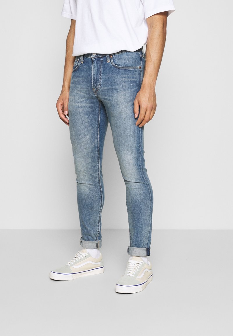 Homme portant un jean slim bleu clair, des chaussettes blanches, des baskets beige et blanc, et un t-shirt blanc sur un fond uni.