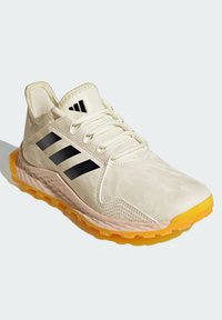 Chaussure de sport de couleur claire avec un dessus texturé, des bandes noires et une semelle extérieure orange vif. Elle dispose d'un renfort à l'avant du pied et d'un système de laçage.