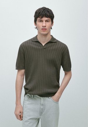 Giovane uomo con capelli corti e scuri che indossa una polo a maniche corte in maglia verde scuro e jeans chiari, in piedi contro uno sfondo chiaro e semplice.