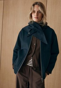 Giacca blu navy scuro con cerniera, collo e due tasche; indossata sopra una camicia marrone e una maglia a righe. Il tessuto sembra morbido e strutturato.