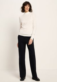 Frau in einem weißen Rollkragenpullover, schwarzen weit geschnittenen Hosen und schwarzen Schuhen, die vor einem hellen Hintergrund steht.