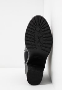 Semelle d’une botte noire avec des crampons à motifs, inclinée contre un coin blanc montrant un design de grip détaillé.