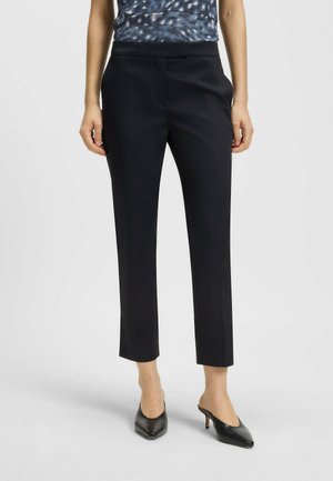 Black tailored pants with a slim fit, smooth fabric, mid-rise waist, and front pleats, paired with pointed black heels and a patterned top.