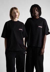 Čierne oversized tričká vyrobené z bavlny, s malým červeným logom na ľavej strane. Obidvaja modeli nosia zodpovedajúce čierne nohavice.