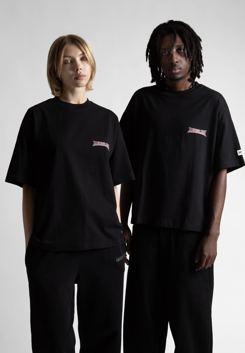 Čierne oversized tričká vyrobené z bavlny, s malým červeným logom na ľavej strane. Obidvaja modeli nosia zodpovedajúce čierne nohavice.