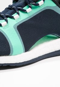 Scarpa sportiva con tomaia in mesh blu navy e verde acqua, dotata di suola bianca ammortizzata. Sistema di allacciatura distintivo e dettagli testurizzati.