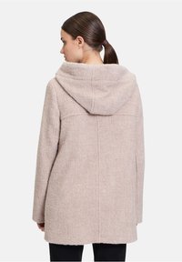 Cappotto con cappuccio in tessuto morbido beige chiaro con finitura testurizzata. Presenta un taglio dritto e dettagli di cuciture visibili sul retro.