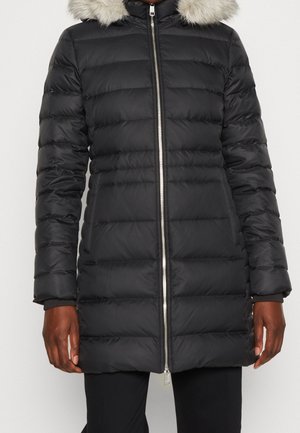 Czarna pikowana zimowa kurtka do kolan z przednim zamkiem błyskawicznym i jasnoszarym kapturem z sztucznego futra, noszona przez osobę o ciemnej karnacji i czarnych spodniach.