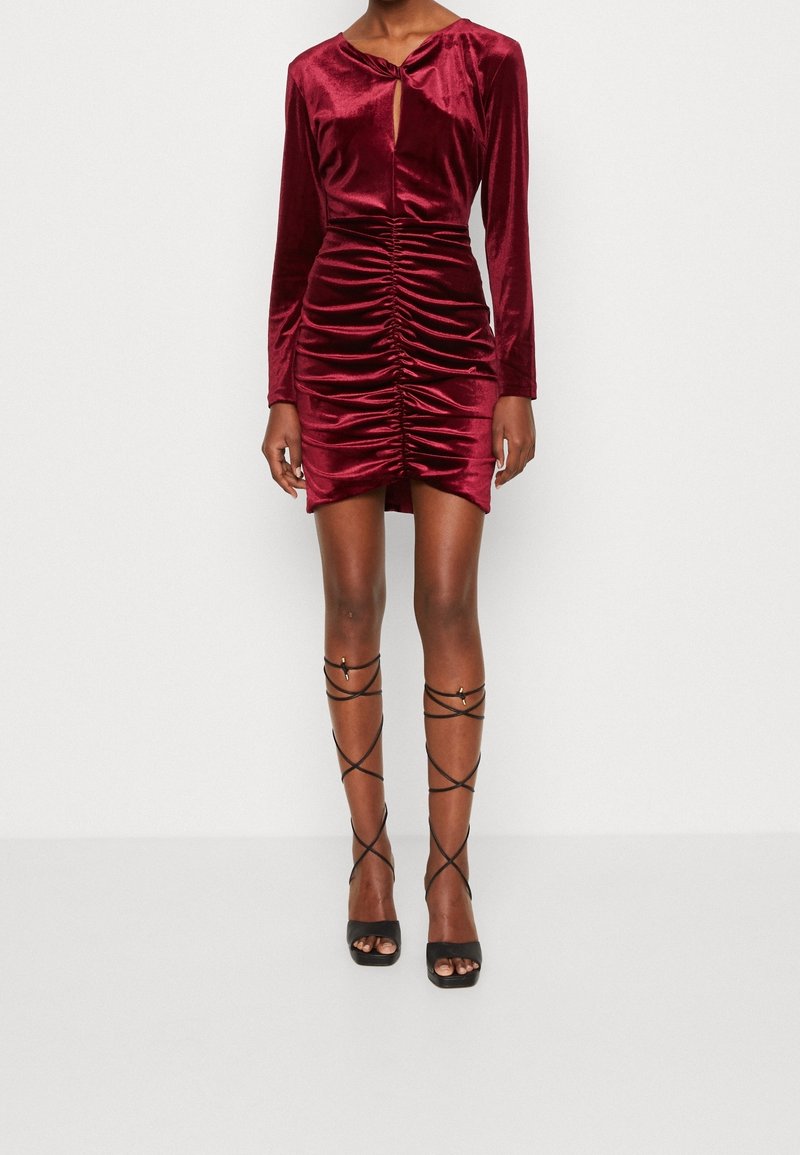Personne portant une mini-robe en velours bordeaux à manches longues froncées et des sandales noires à talons hauts à brides, debout devant un arrière-plan blanc.