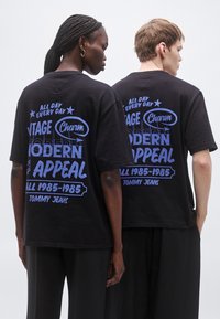 Čierne bavlnené nadrozmerné tričko s modrou grafickou potlačou na zadnej strane. Dizajn obsahuje text a hviezdy, krátke rukávy a okrúhly výstrih.