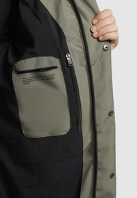 Interiorul unei jachete de culoare verde măslinie și neagră, cu un buzunar cu fermoar și o cusătură expusă, având un mic detaliu de text imprimat. Textură moale.