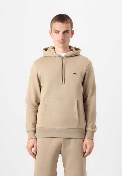 Sweat à capuche beige avec cordon de serrage et poche kangourou. Présente un petit logo vert sur la poitrine. Tissu en mélange de coton doux, coupe décontractée.