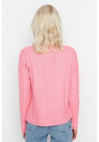 Pull en maille rose avec une coupe décontractée, présentant des détails de couture côtelée et un dos légèrement court. Associé à un jean délavé.