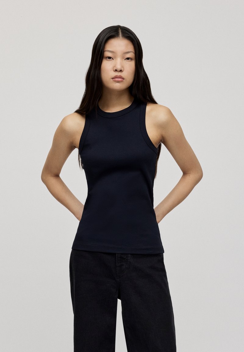 Navy sleeveless top with a high neckline and fitted design. Made of smooth fabric, it features a simple, clean silhouette. Paired with dark jeans.