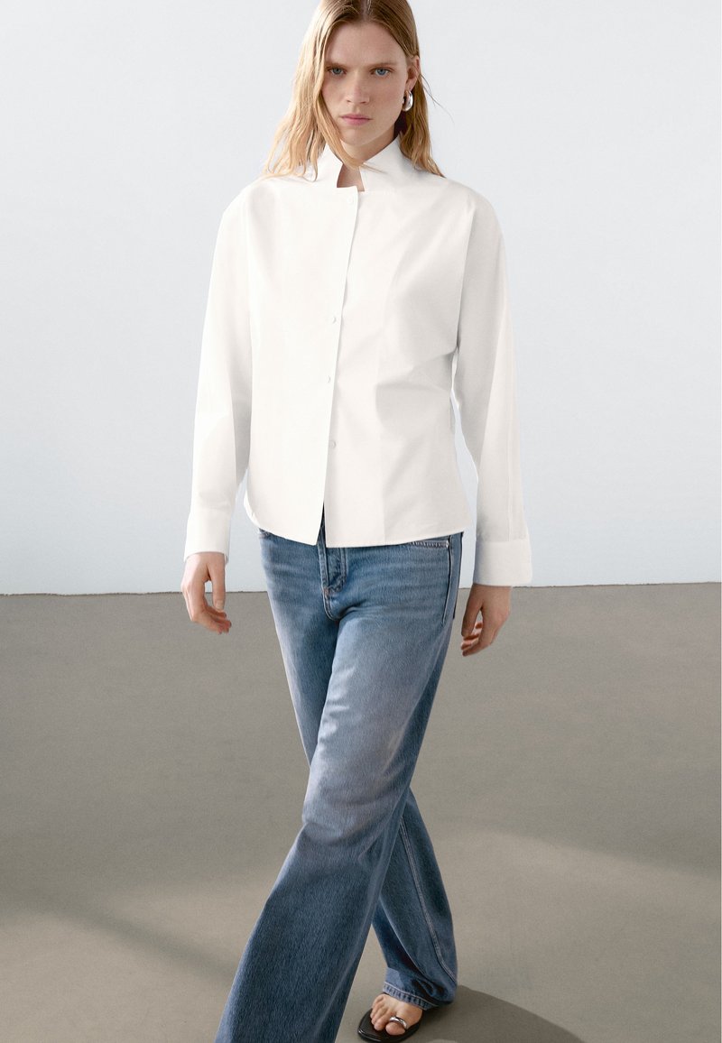 White button-up shirt with a mandarin collar, paired with light blue flared jeans and sandals. Clean lines, smooth fabric texture.