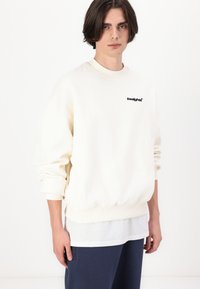 Cream-colored sweatshirt with ribbed cuffs and hem, featuring a small black logo on the chest. Model wears it over a white shirt.