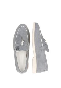 Graue Wildleder-Slipper mit weißer Umrandung, mit dekorativem Tassel und einem Logo auf dem Spann. Weiche Textur, flache Gummisohle.