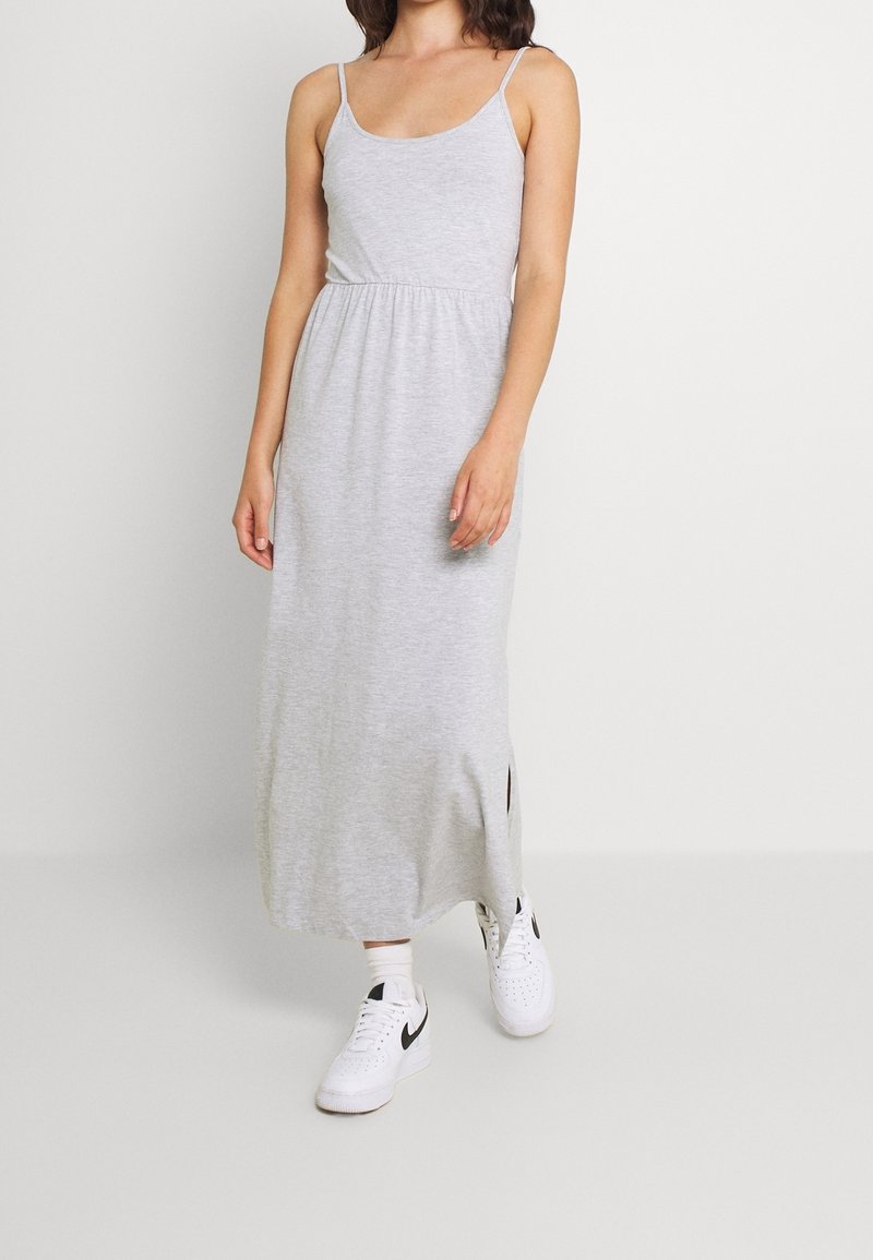 Femme portant une robe longue sans manches gris clair avec des fentes latérales et des baskets blanches, debout devant un fond clair uni.