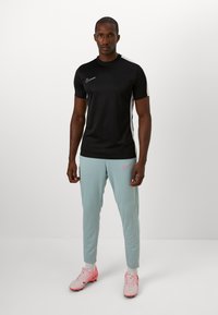 Sort kortærmet Nike-shirt med hvide accenter, parret med lysegrønne Nike joggers og lyserøde fodboldsko, stående på en ensfarvet baggrund.