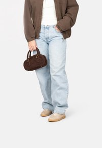 Bolso de ante marrón con dos asas cortas, combinado con un pantalón vaquero suelto de color azul claro y zapatillas beige. La ropa incluye una chaqueta marrón.