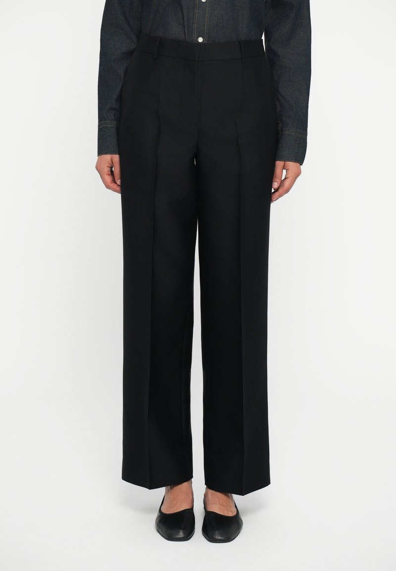 Op maat gemaakte zwarte broek met een hoge taille, wijde pijpen, plooien aan de voorkant en minimale stikseldetails, gedragen met zwarte platte schoenen.
