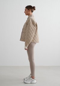 Beige quiltet jakke med en avslappet passform, sidelommer, kombinert med beige leggings og hvite joggesko, presentert mot en ensfarget bakgrunn.