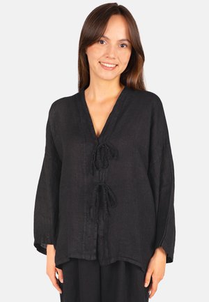 Junge Frau lächelt und trägt ein lockeres schwarzes Langarmshirt mit zwei vorderen Binderverschlüssen vor einem schlichten Hintergrund.