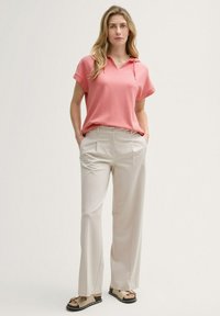 Korallenpinkes kurzärmeliges Oberteil mit Kapuze, kombiniert mit cremefarbenen weiten Hosen. Das Model steht mit den Händen in den Taschen und trägt flache Sandalen.