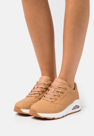Baskets Skechers couleur sable avec semelles intermédiaires blanches et coussin d'air visible portées aux pieds sur un fond clair uni.